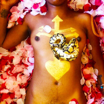 A photo of a nude person laying in a bed of rose petals. They are holding an apple in one hand, have a snake laying on their chest, a crown of flowers on their head, and painted markings on their chest and eye lids.