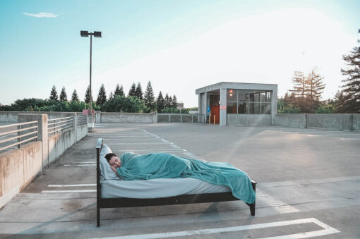 A close up photo of a person sleeping in a bed which is sitting in the middle of an empty parking lot.