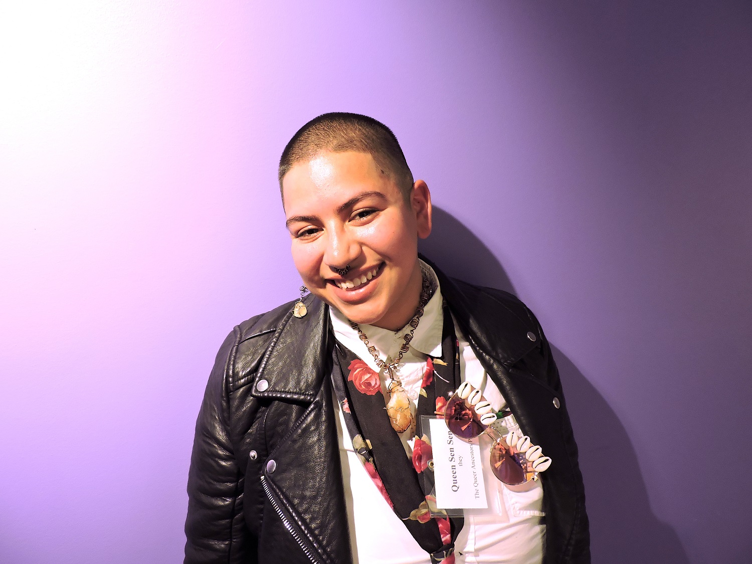 A photo of Queen Sen Sen in front of a purple wall. Sen has short hair. Sen is wearing a black leather jacket and is smiling.