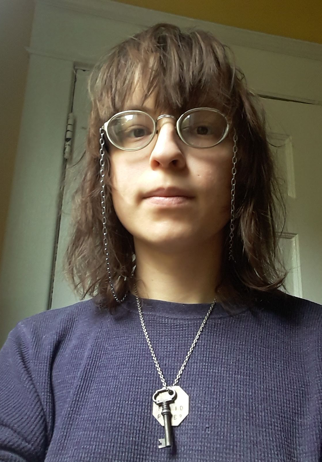 A photo of Z Akhmetova in front of a door. They are wearing a purple shirt, silver glasses, and a silver necklace.